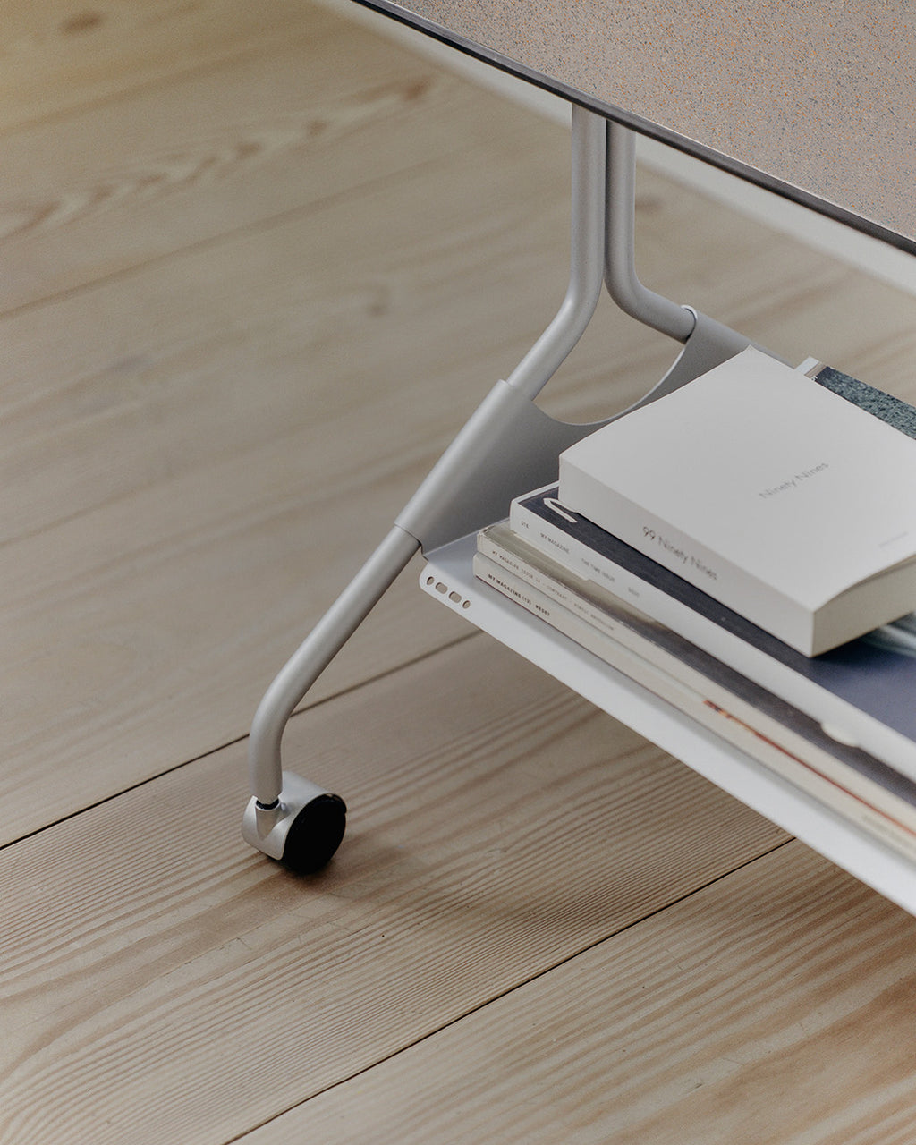 Close-up of Moon in Cloudy Silver stand's curved leg and black caster wheel, with books stacked on the lower shelf
